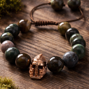 12mm natural agate stone bead bracelet with adjustable brown cord and rose gold tone Viking helmet centerpiece, styled on rustic wood with moss accents in natural warm lighting.